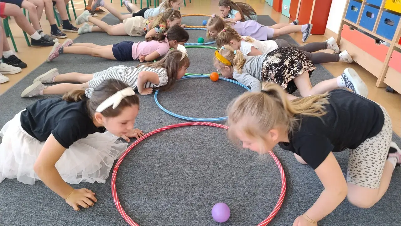Dzieci leżą na podłodze, próbując przesunąć piłeczki za pomocą hula hop. To ciekawe wyzwanie hula hop!