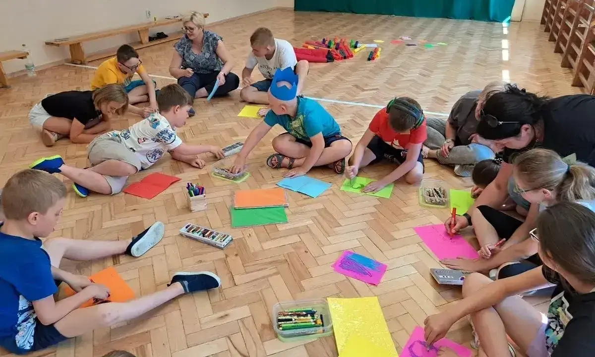 Dzieci i dorośli siedzą na podłodze, rysując i malując. To świetne pomysły na integrację zespołu klasowego, rozwijające kreatywność i współpracę.