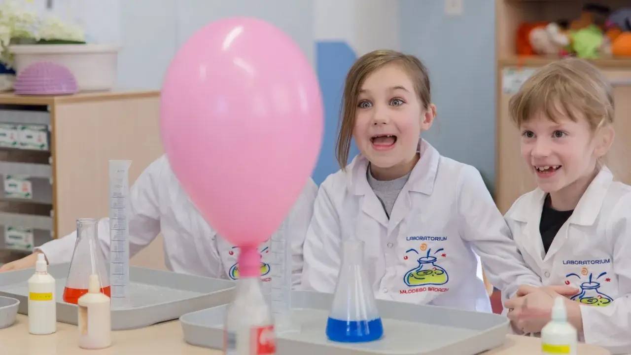 Dzieci w fartuchach laboratoryjnych z ekscytacją obserwują dmuchany balon podczas eksperymentu. To świetna chemia dla dzieci!