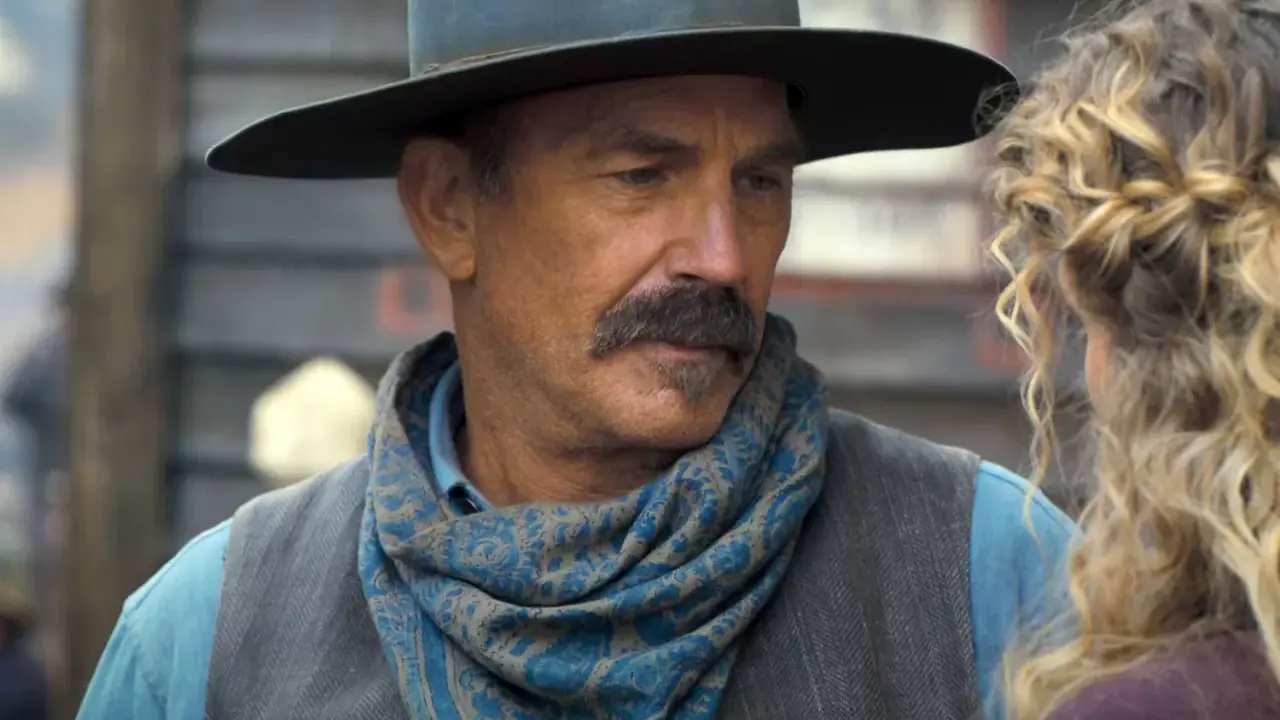 Kevin Costner w filmie z westernowym kapeluszem i chustą na szyi, rozmawia z kobietą.