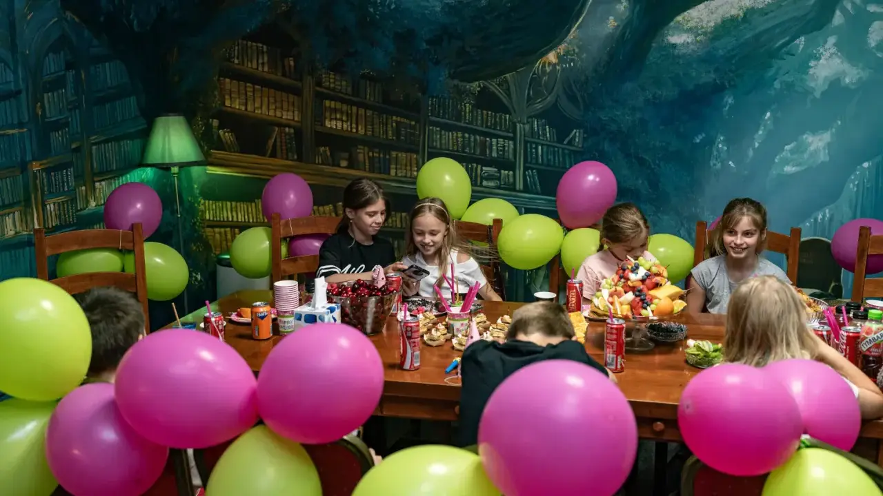 Dzieci świętują urodziny w pokoju zagadek. Kolorowe balony i owocowy tort tworzą radosną atmosferę.