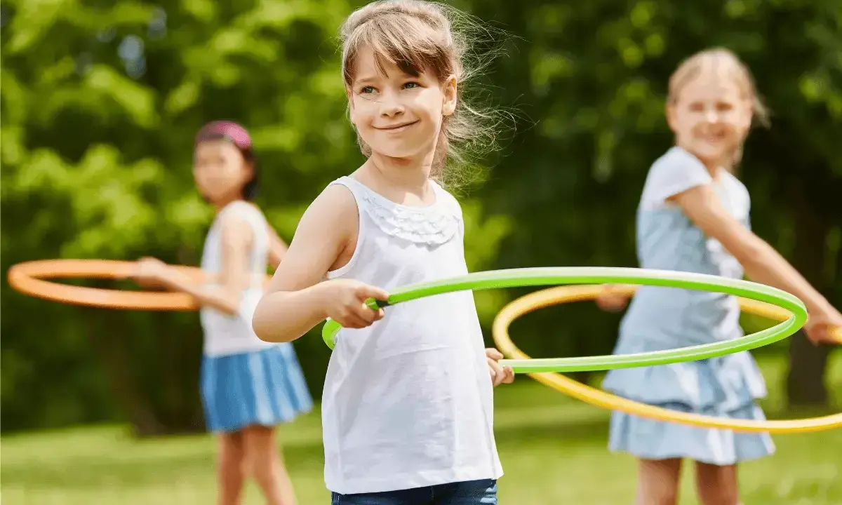 Uśmiechnięte dziewczynki biorą udział w konkurencjach sportowych dla dzieci, kręcąc hula-hop na trawie.
