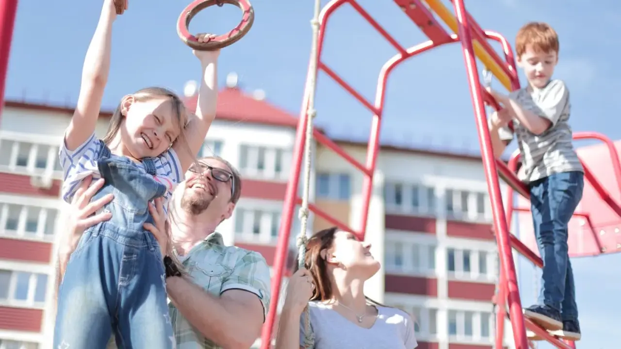 Radosna rodzina na placu zabaw. Ojciec podtrzymuje córkę na kółkach gimnastycznych, syn wspina się po drabince. Zabawy na placu zabaw cele to radość i wspólne chwile.