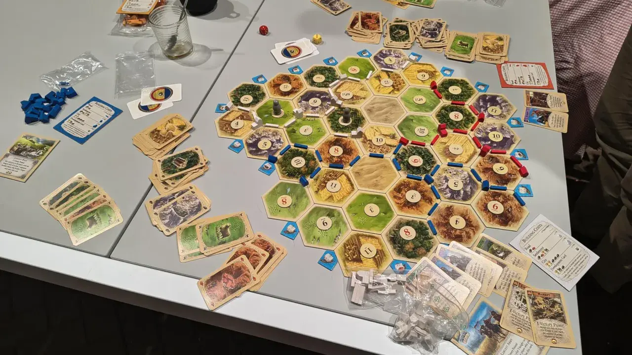 Plansza do gry Catan, karty surowców i rozwoju, pionki. Gracze zastanawiają się nad kolejnością budowania, by zdobyć jak najwięcej punktów.