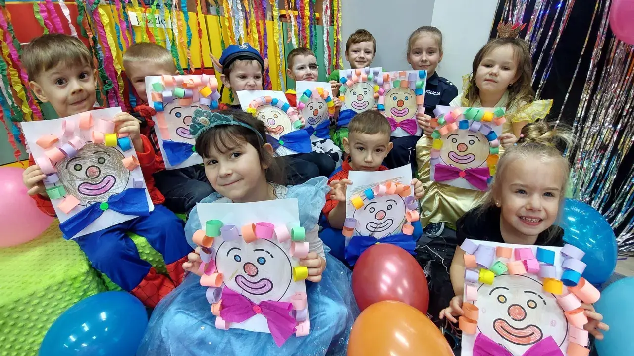 Dzieci w przedszkolu cieszą się z zabaw na balu karnawałowym, prezentując własnoręcznie wykonane prace z wizerunkami klaunów.