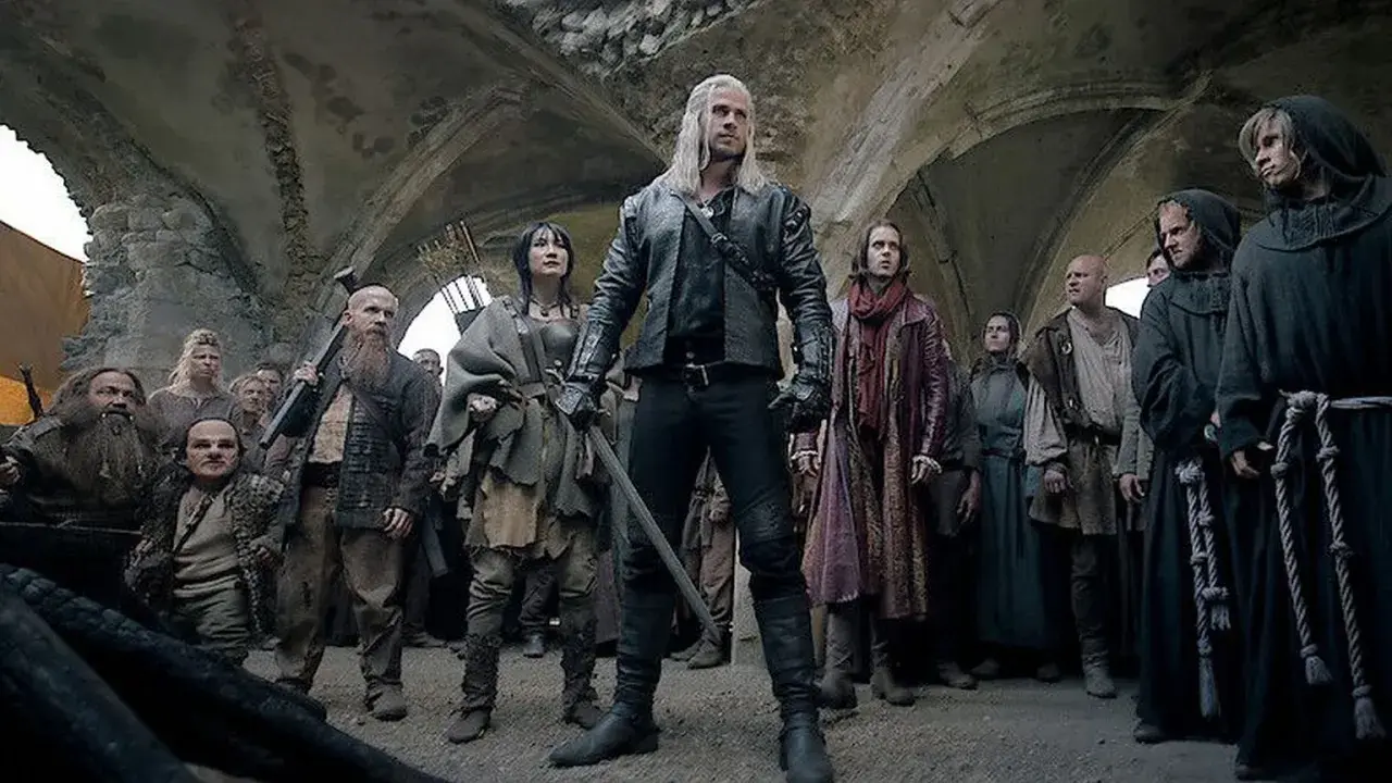 Geralt z Rivii stoi pośród tłumu, gdy ścieżki przeznaczenia w Wiedźminie splatają się. Widać krasnoludy, ludzi i zakapturzonych mnichów.