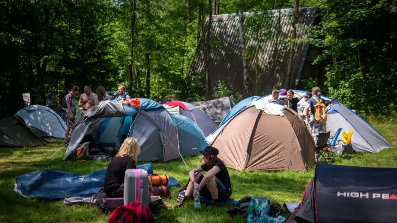 Ludzie rozbijają namioty na polu namiotowym, gotowi na festiwal. W tle widać budynek, a wokół drzewa. To idealne miejsce na nocleg podczas Pyrkonu, gdzie każdy znajdzie swoje miejsce i pyrkon tokeny.