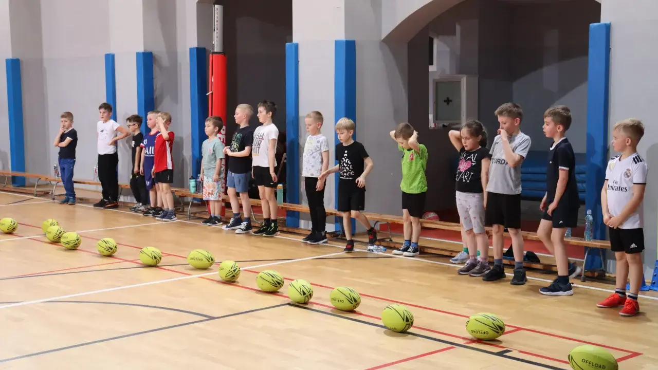 Dzieci w sali gimnastycznej czekają na rozpoczęcie zabaw ruchowych. Przed nimi leżą żółte piłki.