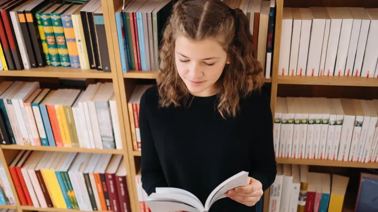 Dziewczynka z warkoczykami czyta książkę na tle regałów pełnych literatury. Może to być streszczenie "Skarbczyka" lub innej lektury.