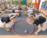 Dzieci leżą na podłodze, próbując przesunąć piłeczki za pomocą hula hop. To ciekawe wyzwanie hula hop!