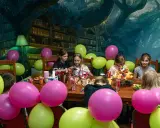 Dzieci świętują urodziny w pokoju zagadek. Kolorowe balony i owocowy tort tworzą radosną atmosferę.