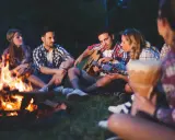 Przyjemne zabawy przy ognisku. Grupa przyjaciół siedzi na trawie, jeden gra na gitarze, inni słuchają.