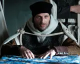 Konrad Wallenrod w zbroi i czarnej czapce, z rękami na stole, otoczony przez rycerzy w białych płaszczach z krzyżami.