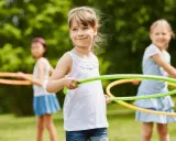 Uśmiechnięte dziewczynki biorą udział w konkurencjach sportowych dla dzieci, kręcąc hula-hop na trawie.