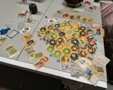 Plansza do gry Catan, karty surowców i rozwoju, pionki. Gracze zastanawiają się nad kolejnością budowania, by zdobyć jak najwięcej punktów.
