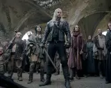 Geralt z Rivii stoi pośród tłumu, gdy ścieżki przeznaczenia w Wiedźminie splatają się. Widać krasnoludy, ludzi i zakapturzonych mnichów.
