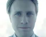 Mężczyzna o niebieskich oczach, z lekko uśmiechniętymi ustami, wygląda jakby był pracownikiem evil corp.
