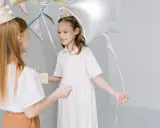 Dwie dziewczynki w czapkach urodzinowych, jedna z prezentem, druga z balonem, przygotowują zabawy na sylwestra dla dzieci.