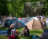 Ludzie rozbijają namioty na polu namiotowym, gotowi na festiwal. W tle widać budynek, a wokół drzewa. To idealne miejsce na nocleg podczas Pyrkonu, gdzie każdy znajdzie swoje miejsce i pyrkon tokeny.