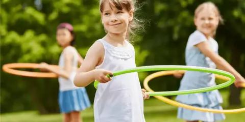 Uśmiechnięte dziewczynki biorą udział w konkurencjach sportowych dla dzieci, kręcąc hula-hop na trawie.
