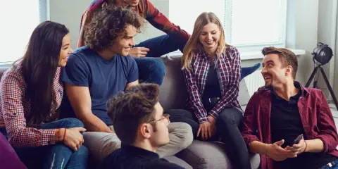 Grupa przyjaciół śmieje się, siedząc na kanapie. Wyglądają, jakby grali w grę, może nawet w Familiadę, odpowiadając na zabawne pytania.