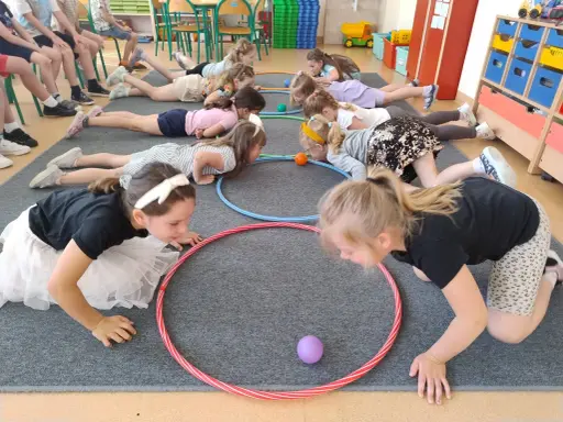 Dzieci leżą na podłodze, próbując przesunąć piłeczki za pomocą hula hop. To ciekawe wyzwanie hula hop!