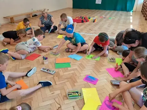 Dzieci i dorośli siedzą na podłodze, rysując i malując. To świetne pomysły na integrację zespołu klasowego, rozwijające kreatywność i współpracę.