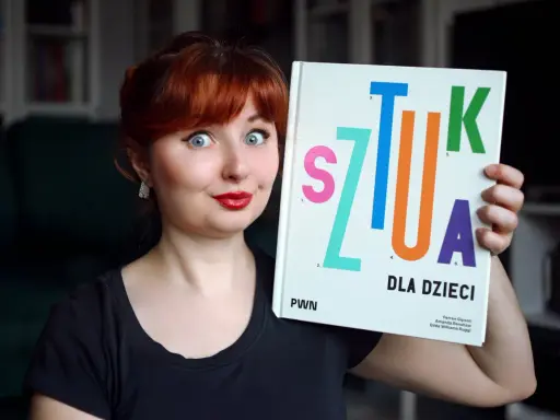 Kobieta z rudymi włosami trzyma książkę "Sztuka dla dzieci". Jej oczy są szeroko otwarte, a usta umalowane na czerwono.