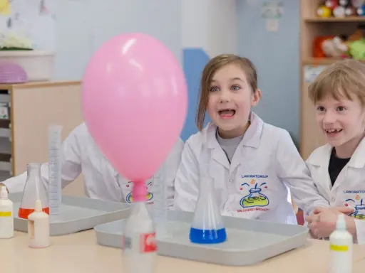 Dzieci w fartuchach laboratoryjnych z ekscytacją obserwują dmuchany balon podczas eksperymentu. To świetna chemia dla dzieci!