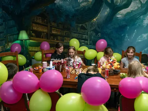 Dzieci świętują urodziny w pokoju zagadek. Kolorowe balony i owocowy tort tworzą radosną atmosferę.