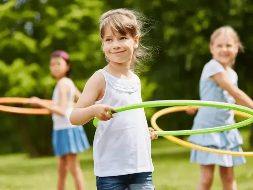 Uśmiechnięte dziewczynki biorą udział w konkurencjach sportowych dla dzieci, kręcąc hula-hop na trawie.