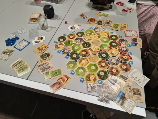 Plansza do gry Catan, karty surowców i rozwoju, pionki. Gracze zastanawiają się nad kolejnością budowania, by zdobyć jak najwięcej punktów.