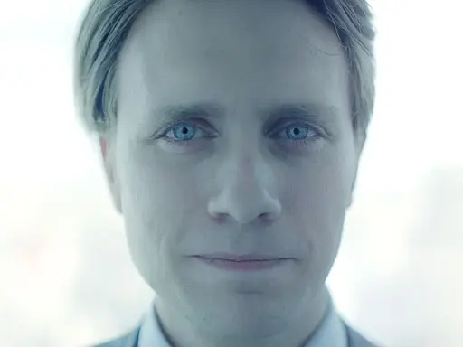 Mężczyzna o niebieskich oczach, z lekko uśmiechniętymi ustami, wygląda jakby był pracownikiem evil corp.