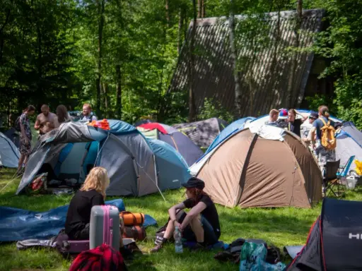 Ludzie rozbijają namioty na polu namiotowym, gotowi na festiwal. W tle widać budynek, a wokół drzewa. To idealne miejsce na nocleg podczas Pyrkonu, gdzie każdy znajdzie swoje miejsce i pyrkon tokeny.