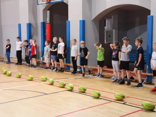 Dzieci w sali gimnastycznej czekają na rozpoczęcie zabaw ruchowych. Przed nimi leżą żółte piłki.