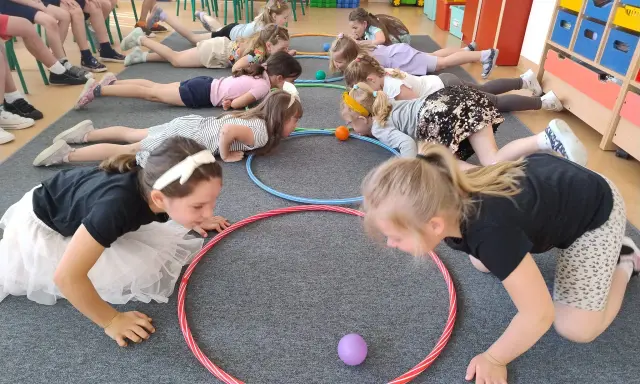 Dzieci leżą na podłodze, próbując przesunąć piłeczki za pomocą hula hop. To ciekawe wyzwanie hula hop!