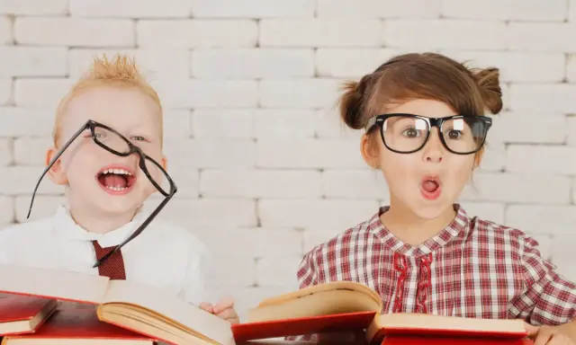 Dzieci w okularach śmieją się i otwierają usta z zaskoczenia, otoczone książkami. Wesołe zabawy w klasie!