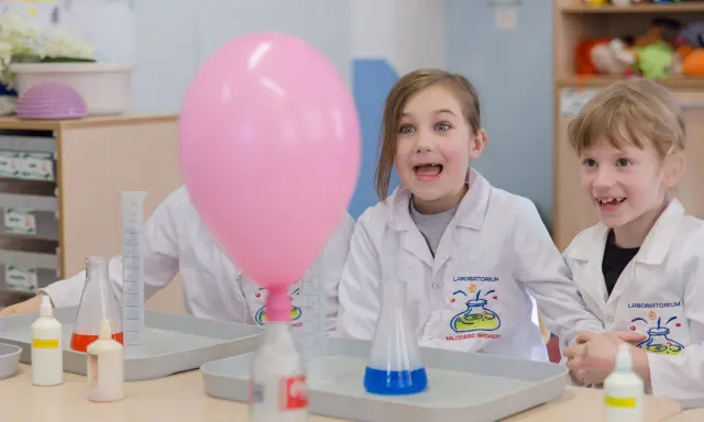 Dzieci w fartuchach laboratoryjnych z ekscytacją obserwują dmuchany balon podczas eksperymentu. To świetna chemia dla dzieci!
