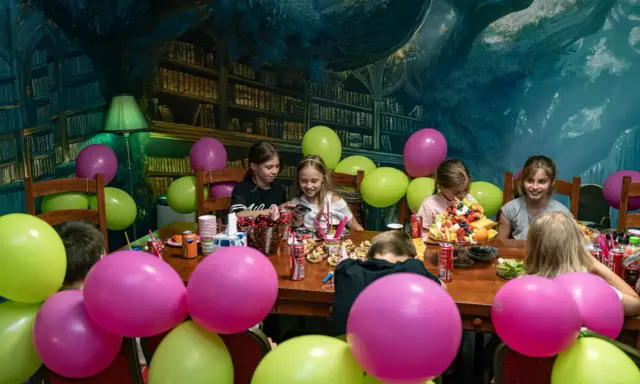 Dzieci świętują urodziny w pokoju zagadek. Kolorowe balony i owocowy tort tworzą radosną atmosferę.