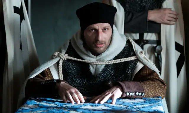 Konrad Wallenrod w zbroi i czarnej czapce, z rękami na stole, otoczony przez rycerzy w białych płaszczach z krzyżami.