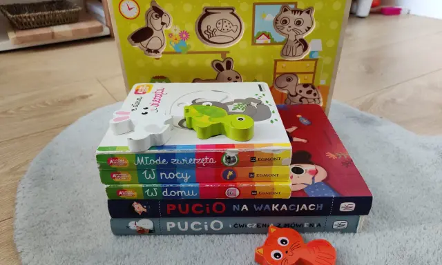 Zabawki dla 1,5 rocznego dziecka: drewniana układanka ze zwierzątkami i stos książeczek "Akademia Mądrego Dziecka" oraz "Pucio".