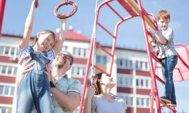 Radosna rodzina na placu zabaw. Ojciec podtrzymuje córkę na kółkach gimnastycznych, syn wspina się po drabince. Zabawy na placu zabaw cele to radość i wspólne chwile.