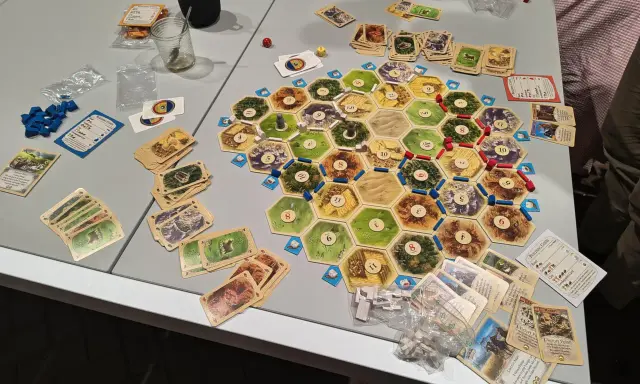 Plansza do gry Catan, karty surowców i rozwoju, pionki. Gracze zastanawiają się nad kolejnością budowania, by zdobyć jak najwięcej punktów.