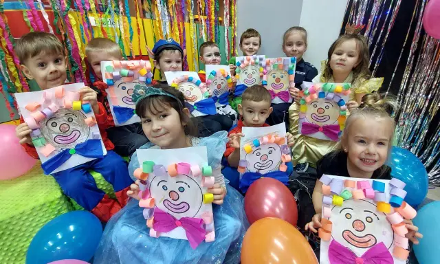 Dzieci w przedszkolu cieszą się z zabaw na balu karnawałowym, prezentując własnoręcznie wykonane prace z wizerunkami klaunów.
