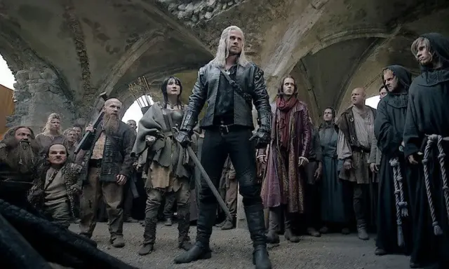 Geralt z Rivii stoi pośród tłumu, gdy ścieżki przeznaczenia w Wiedźminie splatają się. Widać krasnoludy, ludzi i zakapturzonych mnichów.