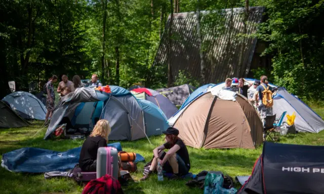 Ludzie rozbijają namioty na polu namiotowym, gotowi na festiwal. W tle widać budynek, a wokół drzewa. To idealne miejsce na nocleg podczas Pyrkonu, gdzie każdy znajdzie swoje miejsce i pyrkon tokeny.