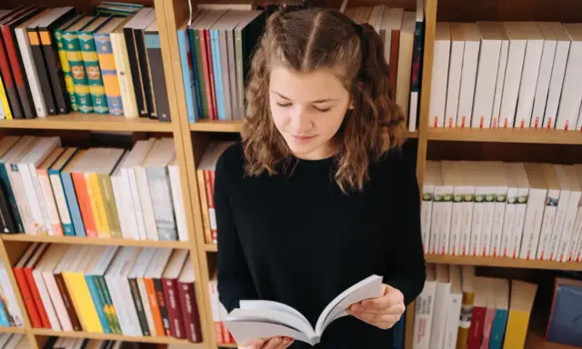 Dziewczynka z warkoczykami czyta książkę na tle regałów pełnych literatury. Może to być streszczenie "Skarbczyka" lub innej lektury.