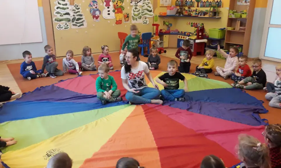 Dzieci i pani bawią się w kole na kolorowym spadochronie. Radosne zabawy w kole dla starszych dzieci w przedszkolu.
