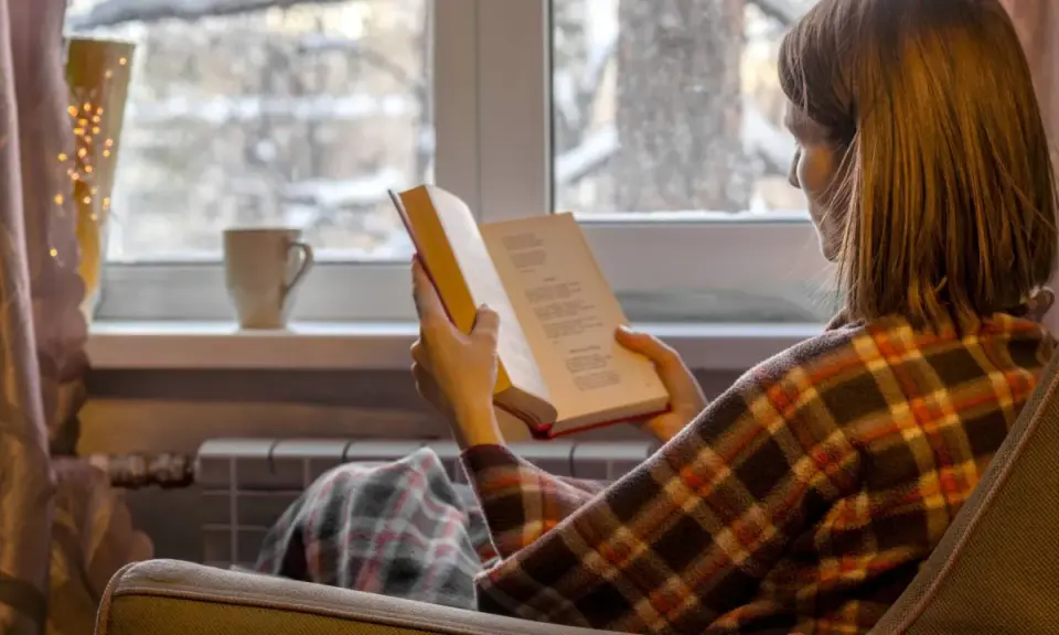 Kobieta w kratę czyta książkę przy oknie, idealne książki na zimowe wieczory.