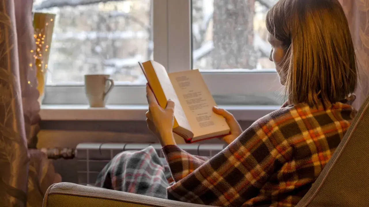 Kobieta w kratę czyta książkę przy oknie, idealne książki na zimowe wieczory.